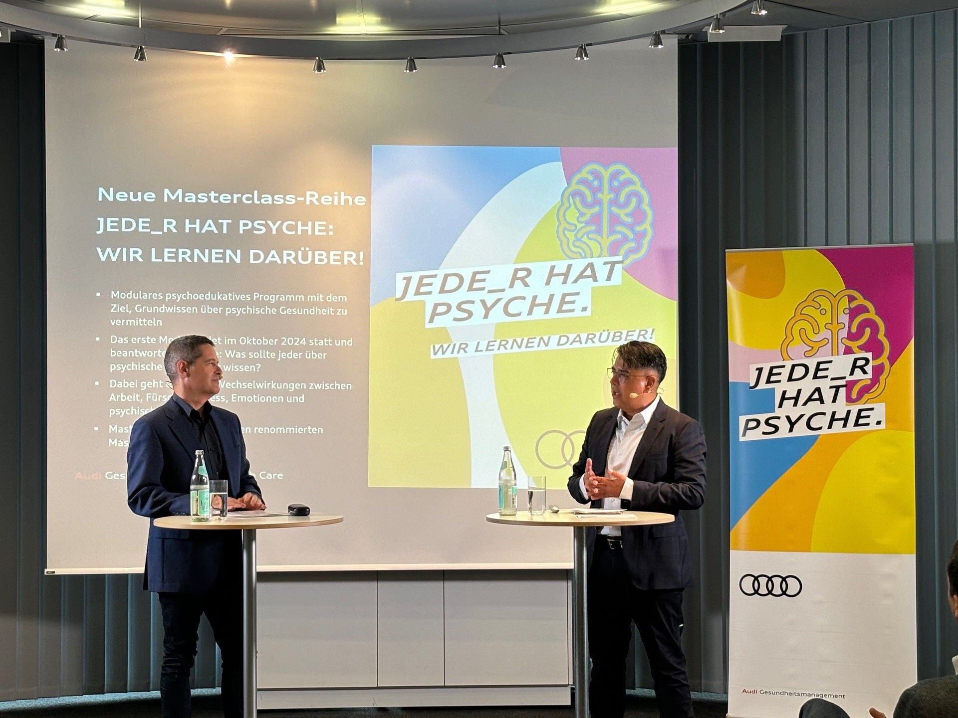 Two men speak on stage at an Audi event for the series “Everyone has a psyche” with banners and a presentation in the background.